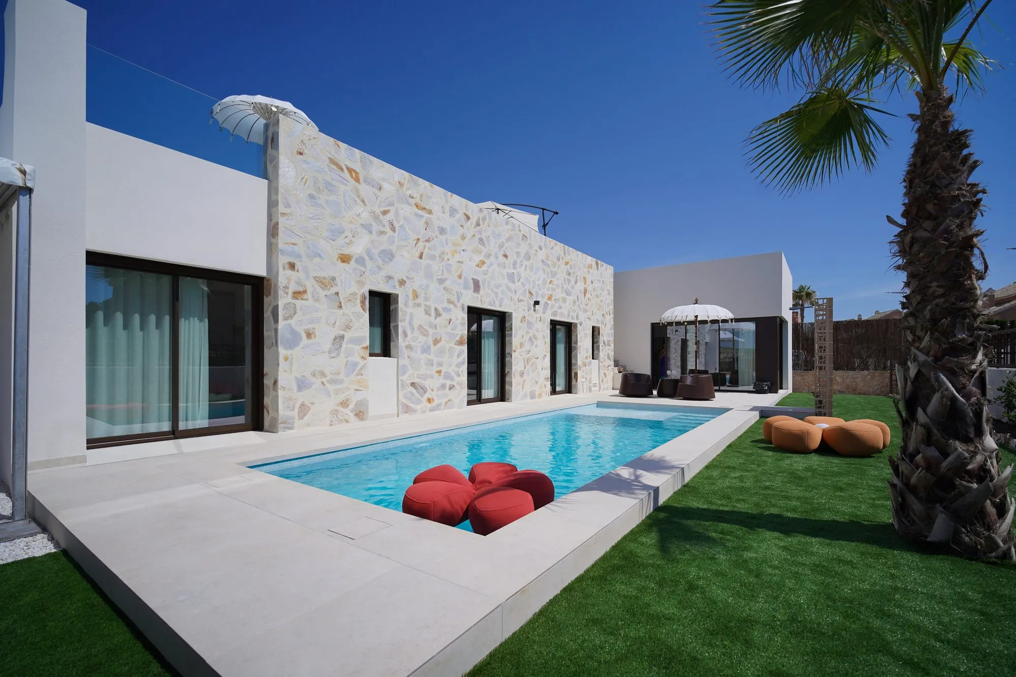 La villa témoin de LIEGE Mediterránea : piscine privée le long de la façade en pierre naturelle, avec pelouse, palmier et parasol