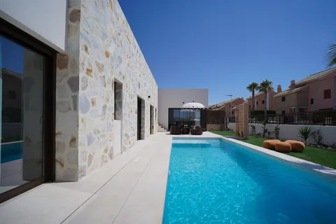 Villa témoin : la piscine le long de la façade en pierre, avec la baie vitrée du séjour à gauche et la pelouse à droite